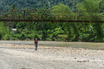 Man standing near the river with old hanging bridge, in Lokop Serbajadi, east Aceh, Indonesia