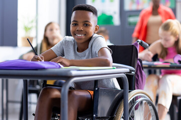 Fototapeta na wymiar Portrait of smiling african american schoolboy in wheelchair working at desk in class