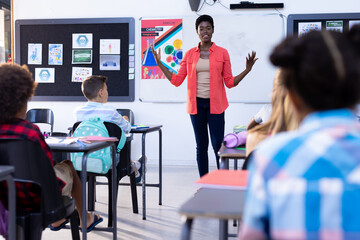 Diverse pupils sitting at desks listening to female teacher at front of class