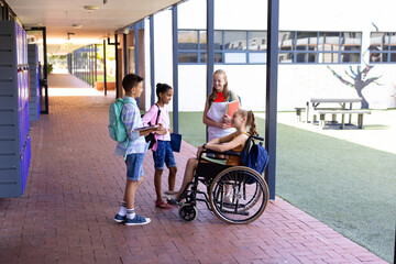 Happy diverse school children talking with girl in wheelchair in school corridor, copy space