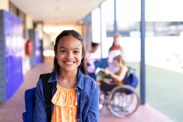 Portrait of smiling biracial schoolgirl standing with bag in school corridor