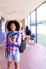 Portrait of smiling biracial schoolgirl standing with bag in school corridor copy space