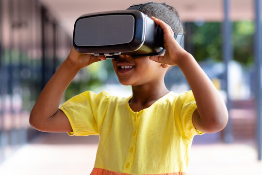 Happy biracial schoolgirl using vr headset in sunny school corridor