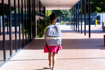 Rear view of biracial schoolgirl with school bag walking in corridor outside school, with copy space
