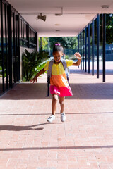 Vertical of happy biracial schoolgirl with school bag, running outside school, with copy space