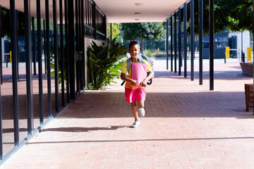 Happy biracial schoolgirl with school bag, holding books and running outside school, with copy space