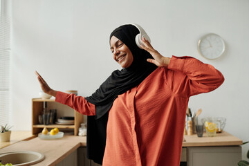 Young cheerful Muslim woman with headphones, hijab and shirt listening to her favorite music,...