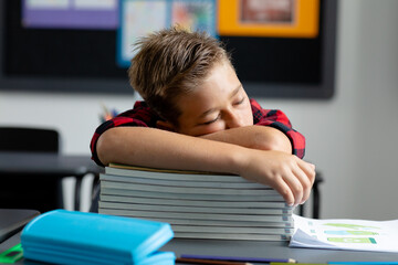 Tired caucasian schoolboy sitting at desk sleeping on pile of books in classroom