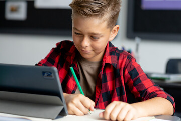 Happy caucasian schoolboy sitting at desk using tablet in class and writing