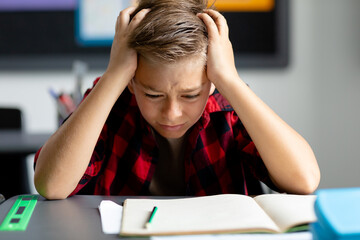 Stressed caucasian schoolboy at desk, head in hands, staring at book in class with copy space