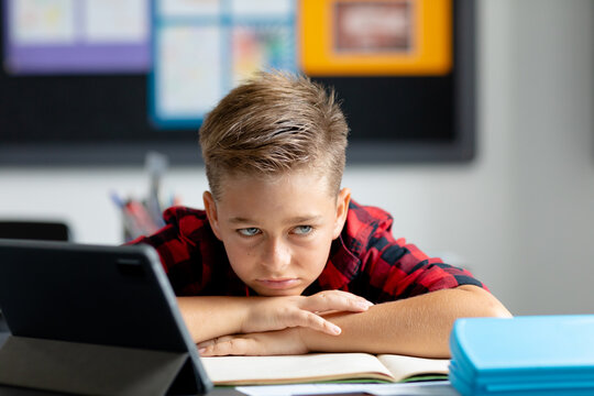 Bored, sad caucasian schoolboy sitting at desk with tablet, leaning on books