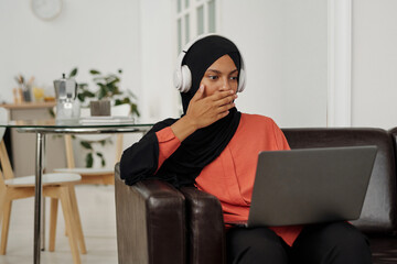 Young shocked Muslim woman in hijab covering her mouth by hand while watching online video or movie on laptop screen at leisure
