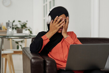 Young frightened Muslim woman with laptop on knees covering her face with hands while sitting on...
