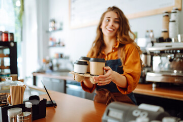Fototapeta premium Best coffee for you. Cheerful woman in an apron at the bar counter holds coffee glasses in a cafe. Takeaway food concept.