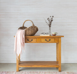 Full length view of oak side table against white wood panelled wall with wicker basket, pink scarf, dried flowers, book and cup (selective focus)