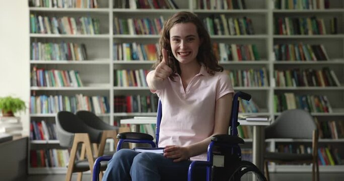 Serious young student with disability using wheelchair, posing in library, holding book, looking at camera, getting cheerful, happy, showing like thumb up hand with toothy smile