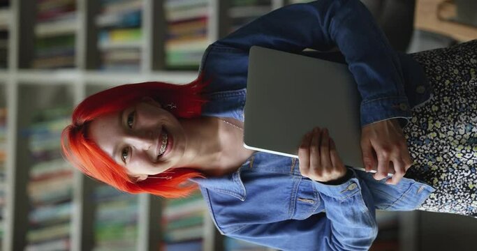 Serious teenage hipster student girl holding tablet, standing in university library, looking at camera, getting happy, cheerful, smiling. Vertical portrait of model with red hair and teeth brackets
