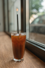 A glass of refreshing coffee with tonic, lemon, ice cubes and straw on wooden background next to the window.
