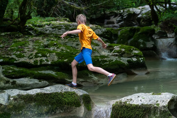 Active boy jumps from rock to rock on a calm mountain river hidden in a dense forest