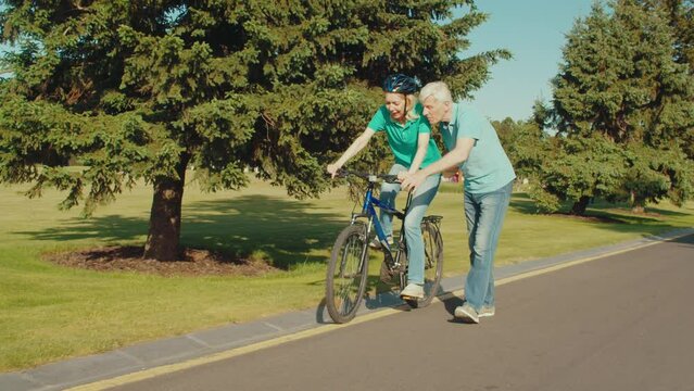 Joyful happy beautiful mature woman in cycling helmet learning to ride bicycle and caring handsome man holding bike and helping to keep balance, while retired couple relaxing in summer park.
