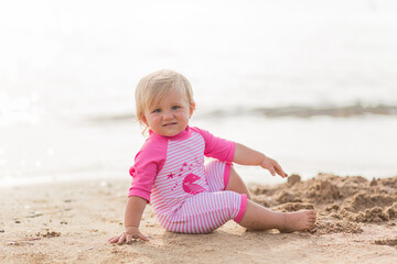 a cute little happy smiling tanned caucasian kid toddler baby girl in one-piece striped pink swimsuit plays in the sand  on the beach. Water safety. Vacation and family holiday 