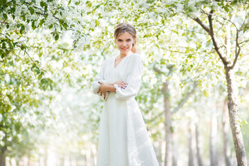 Portrait of a young woman in a white dress against the backdrop of blooming apple trees. The girl poses against the background of flowers in the spring park.