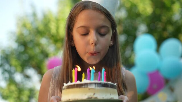 Close-up portrait of happy girl blowing out candles on birthday cake in slow motion smiling. Positive satisfied Caucasian child celebrating holiday outdoors in park on sunny summer day