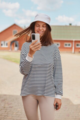 Smiling young beautiful caucasian woman wearing striped shirt and panama using smartphone standing at street having video call or making selfie checking network.