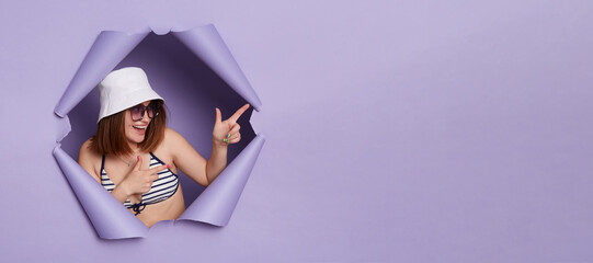 Smiling attractive woman wearing swimwear panama and sunglasses breaking through purple paper background pointing fingers at advertisement area.