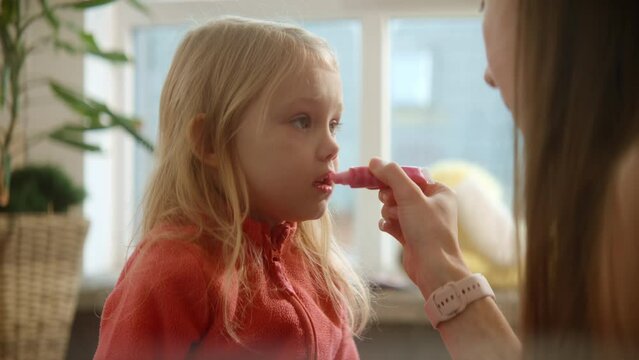 Mother applies lip balm precisely on daughter's lips, beauty care process, girl's growing. Family weekend, female beauty routine, sunny nursery room background. High quality 4k footage