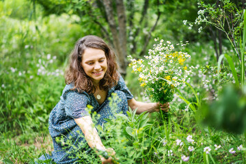 Young cheerful smiling woman in a summer dress picking a bouquet of wild flowers in a summer garden