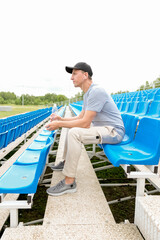 Male sportsman wearing earphones on stadium outside sitting on tribune listening to music holding bottle drinking water