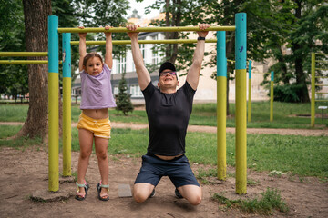 Father with child girl doing pull-ups on workout outdoor area. Healthy active lifestyle, happy family time. Modern fatherhood concept 