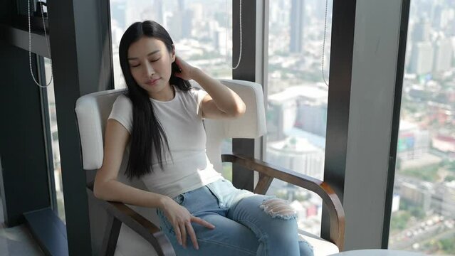 Asian woman sitting by window, enjoying city view, relaxed and contemplative.