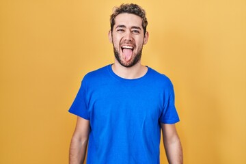 Hispanic man with beard standing over yellow background sticking tongue out happy with funny...