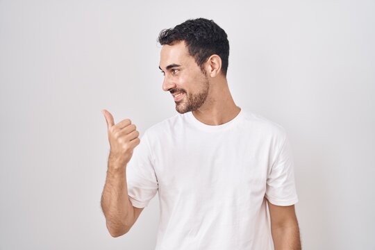 Handsome hispanic man standing over white background smiling with happy face looking and pointing to the side with thumb up.