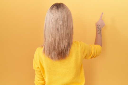 Young caucasian woman wearing yellow sweater posing backwards pointing ahead with finger hand