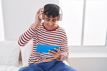 Young hispanic man watching video on touchpad sitting on bed at bedroom