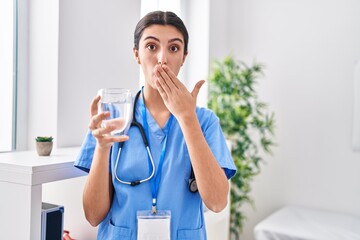 Young beautiful woman wearing doctor uniform and stethoscope drinking water covering mouth with hand, shocked and afraid for mistake. surprised expression