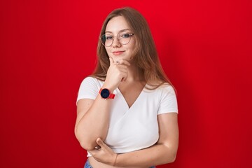 Young caucasian woman standing over red background looking confident at the camera smiling with crossed arms and hand raised on chin. thinking positive.