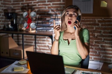 Young beautiful woman working at the office at night speaking on the phone amazed and surprised looking up and pointing with fingers and raised arms.