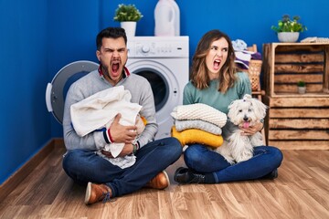 Young hispanic couple doing laundry sitting on the floor angry and mad screaming frustrated and furious, shouting with anger. rage and aggressive concept.