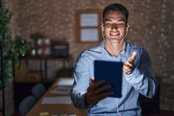 Handsome hispanic man working at the office at night smiling cheerful offering palm hand giving assistance and acceptance.
