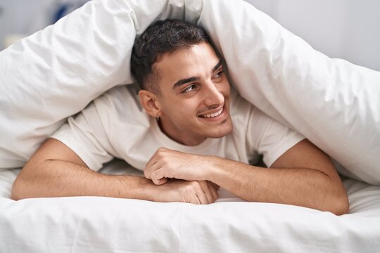 Young hispanic man lying on bed covering with bedsheet at bedroom