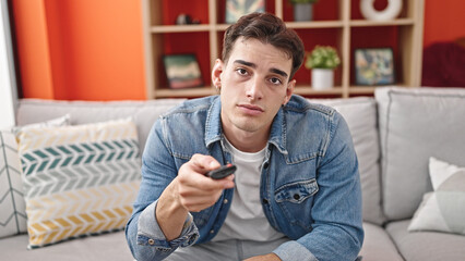 Young hispanic man watching tv sitting on sofa with boring expression at home