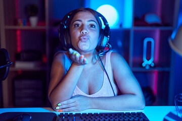 Young blonde woman playing video games wearing headphones looking at the camera blowing a kiss with...