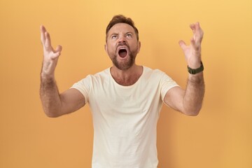 Fototapeta premium Middle age man with beard standing over yellow background crazy and mad shouting and yelling with aggressive expression and arms raised. frustration concept.