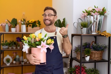 Middle age man with beard working at florist shop holding plant smiling happy and positive, thumb up doing excellent and approval sign