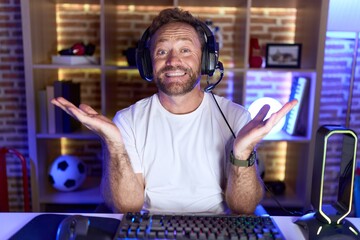 Middle age man with beard playing video games wearing headphones smiling cheerful offering hands giving assistance and acceptance.