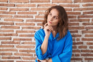 Beautiful brunette woman standing over bricks wall with hand on chin thinking about question, pensive expression. smiling with thoughtful face. doubt concept.